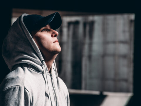 The image shows a young man wearing a hooded grey jumper, looking up as if taking a large sigh.