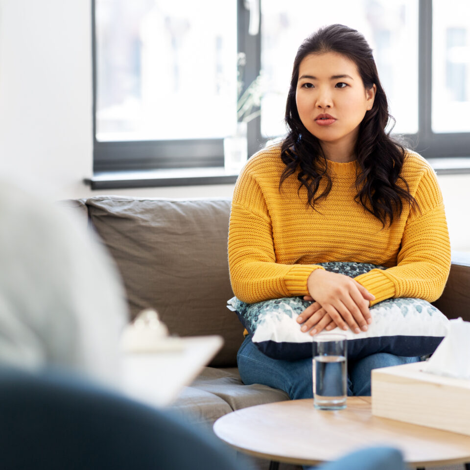 A young lady in a counselling session. She has dark hair and wears a yellow jumper. We only see the counsellor from behind, but they appear to be a woman with dark hair, wearing a white top.