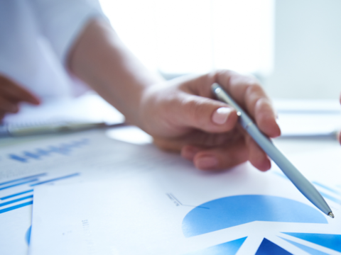The image shows a female hand, holding a pen and skimming over documents that shows various charts and data.